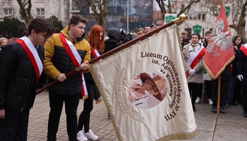Trójka młodych ludzi, w środku chłopiec, który trzyma pochylony w hołdzie sztandar XVII Liceum Ogólnokształcącego im. Agnieszki Osieckiej we Wrocławiu 