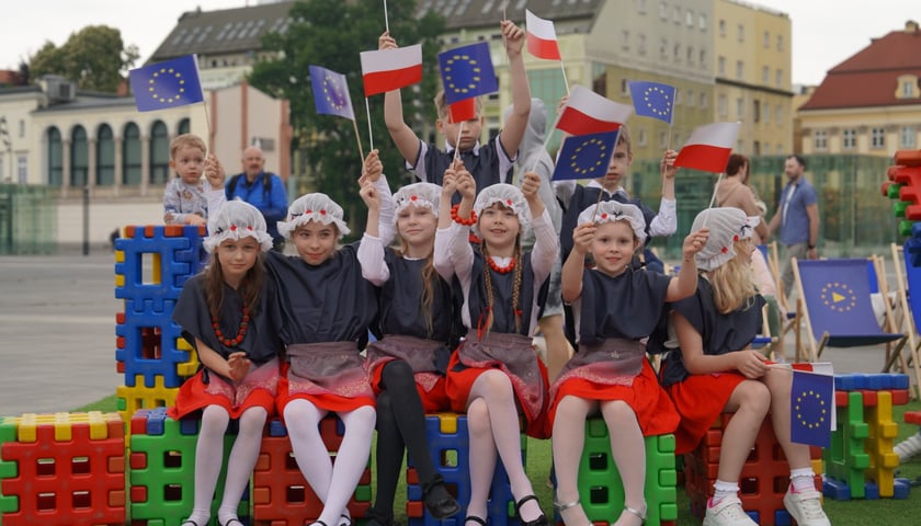Grupa dziewczynek w strojach ludowych siedząca na wielkich klockach i trzymająca chorągiewki Polski i Unii Europejskiej. Dzień Europy na placu Wolności we Wrocławiu, 11.05.2024