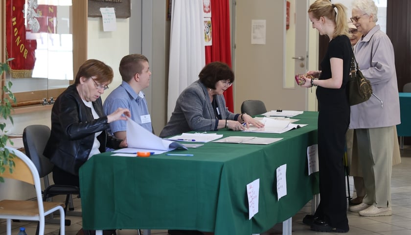 Wybory we Wrocławiu. Zdjęcie ilustracyjne. Na zdjęciu kilka osób stoi w kolejce przed stołem, za którym siedzą kobiety i sprawdzając listy wyborcze