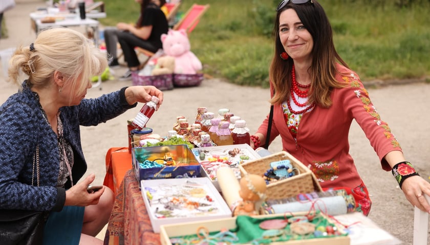 Kobieta podnosi butelkę z płynem ze stoiska na pchlim targu. Za stołem siedzi uśmiechnięta handlarka