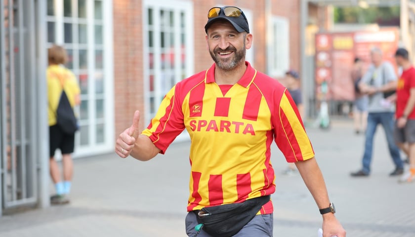 Uśmiechnięty mężczyzna, unoszący kciuk, w żółto-czerwonej koszulce Sparty przed stadionem 