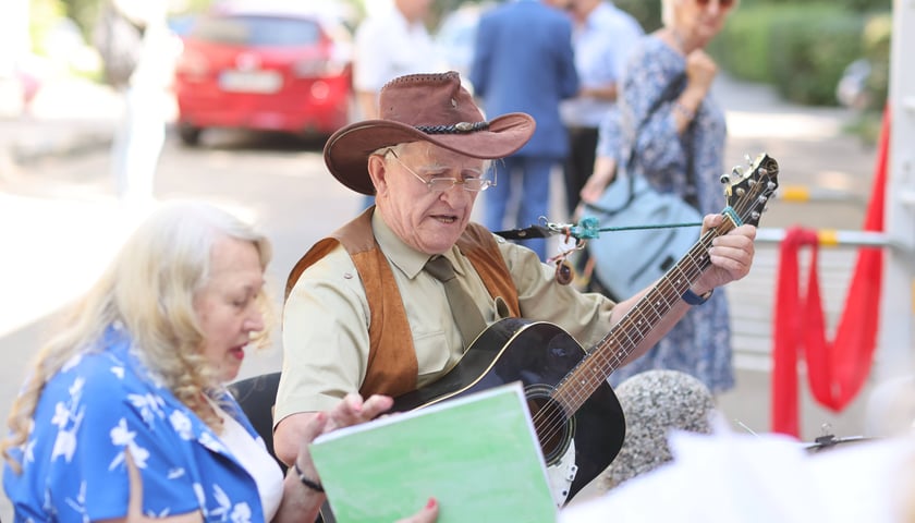 Mężczyzna w kapeluszu z szerokim rondem gra na gitarze, obok śpiewa kobieta w niebieskiej sukni. Otwarcie Centrum Aktywności Lokalnej na Hubach (31.07.2024)