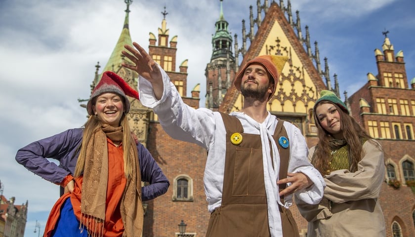 Troje ludzi przebranych w kolorowe krasnalowe stroje na tle wrocławskiego ratusza 