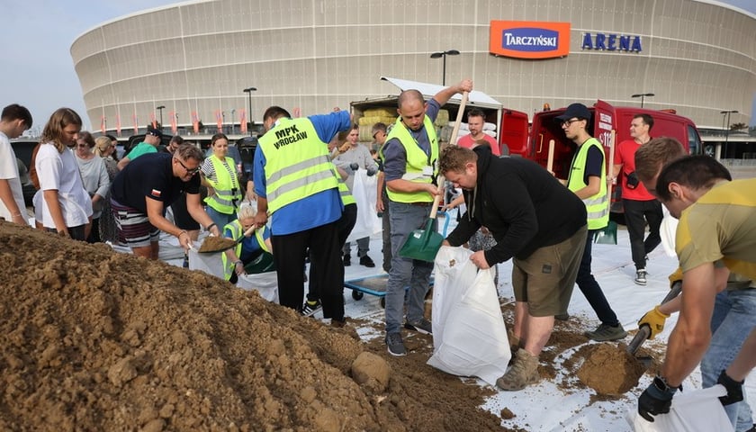 Grupa osób wokół hałdy piachu, sypią go do worków, za nimi widać stadion