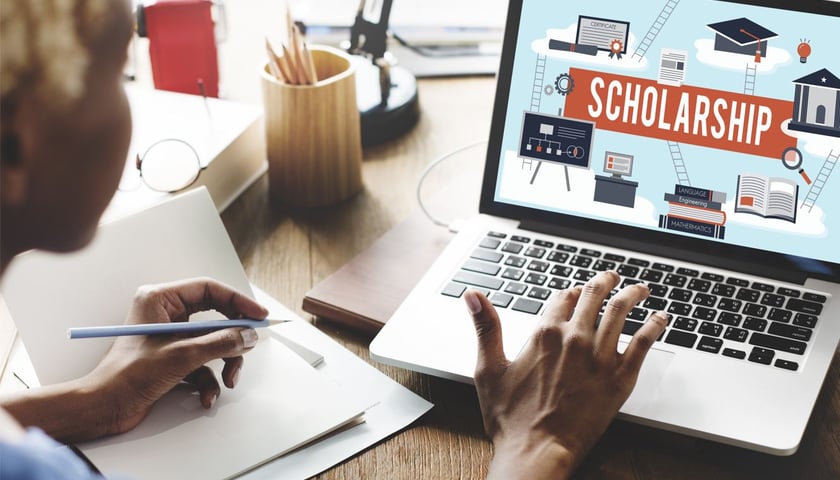 Kobieta siedzi przy biurku, w ręce trzyma ołówek i patrzy na ekran laptopa