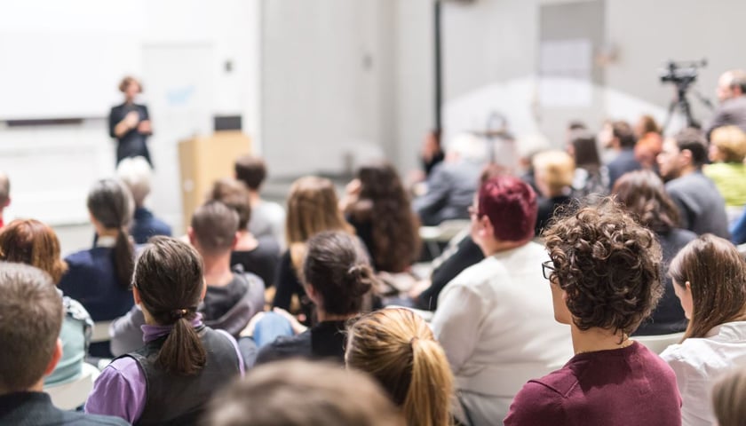 Sala wykładowa pełna studentek i studentów. W tle prowadzący zajęcia