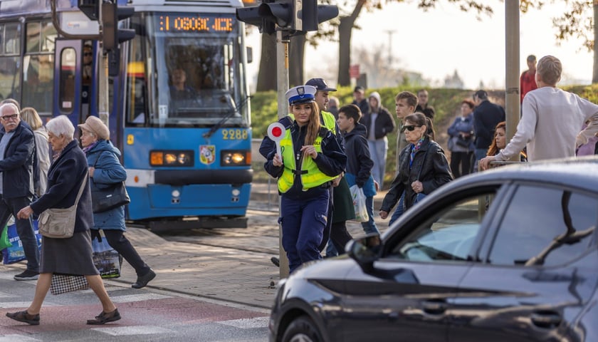 Okolice cmentarza Osobowickiego w dniu Wszystkich Świętych. Widać tramwaj, przechodniów, policjantkę i samochód
