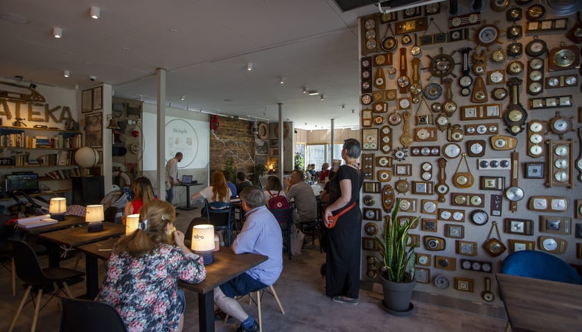 Na pierwszym planie kolekcja barometrów, na drugim ludzie siedzący przy stolikach słuchają wykładu, w tle wykładowca przy obrazie z rzutnika