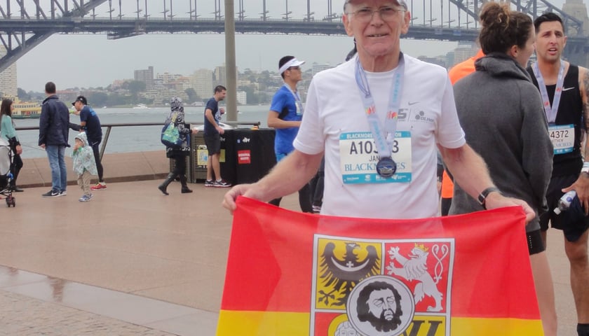 Biegacz na zdjęciu w stroju sportowym to prof. Jan Chmura. Trzyma czerwono-żółtą flagę z herbem Wrocławia, którą zawsze zabiera ze sobą na maratony. Zdjęcie zrobione po zakończeniu Blackmores Sydeny Marathon na tle słynnego Harbour Bridge. Wrocławianin startował w ramach Korony 7 Kontynentów. Zajął 3. miejsce w kategorii 65-69