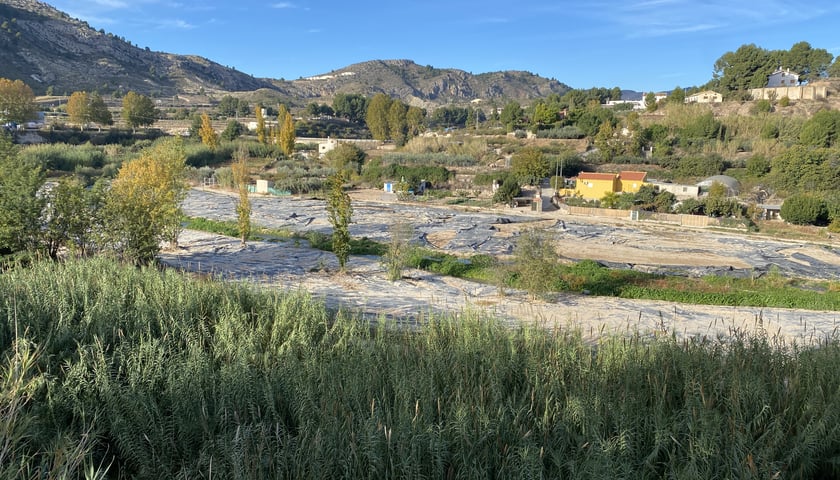 Na zdjęciu obszerny krajobraz pokazujący dolinę z małymi budynkami. Na skarpie zagospodarowane przestrzenie pól. W tyle pobliskie pasmo górskie i niebo. /The photo shows a vast landscape showing a valley with small buildings. On the slope are cultivated fields. In the background are the nearby mountain range and the sky.