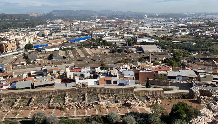 Na zdjęciu widoczne z lotu ptaka miasto z budynkami mieszkalnymi i przemysłowymi. Za miastem widoczne góry i niebo z chmurami./ The photo shows a city with residential and industrial buildings visible from from above. Beyond the city are mountains and a cloudy sky.
