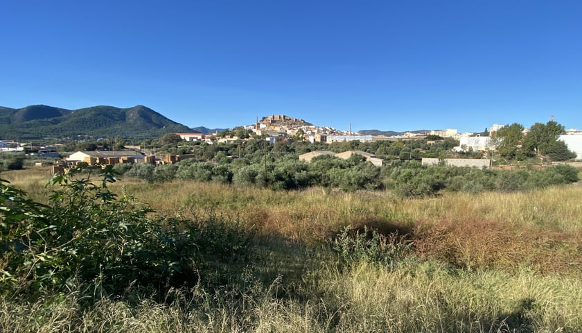 Na zdjęciu widoczny pagórkowaty obszar pokryty niską roślinnością. Za nią znajdują się budynki mieszkalne i przemysłowe. W tle góry i niebo./ The photo shows a hilly area covered with low vegetation. Behind it are residential and industrial buildings. In the background are mountains and sky.