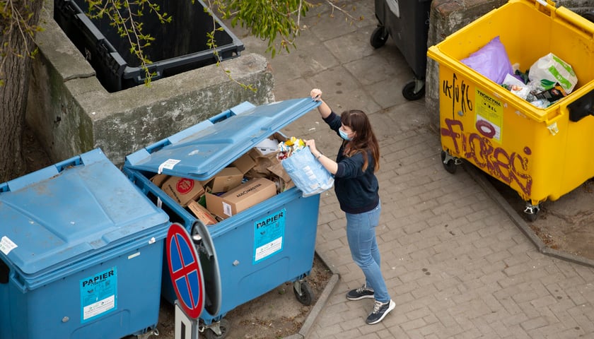 Powiększ obraz: Młoda kobieta wyrzuca kartonowe opakowania do niebieskiego kontenera na śmieci z napisem PAPIER