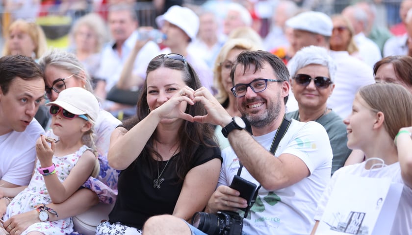 Publiczność siedząca na krzesełkach w Rynku na Old Jazz Days. Na pierwszym planie kobieta i mężczyzna w okularach, uśmiechają się i układają dłonie w kształt serca