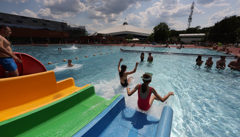 Powiększ obraz: <p>Letnie kąpielisko Orbita przy Wejherowskiej. Dzieci wskakują do wody z ż&oacute;łto-zielono-niebieskiej zjeżdżalni, z lewej mężczyzna w niebieskich szortach, w basenie stoi kilka os&oacute;b</p>