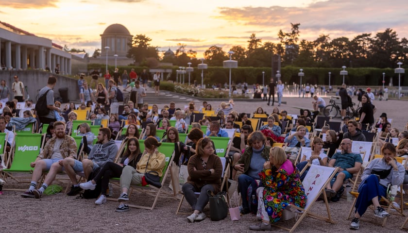 Siedzący ludzie pod Halą Stulecia podczas letniego kina odbywającego się po zachodzie słońca 
