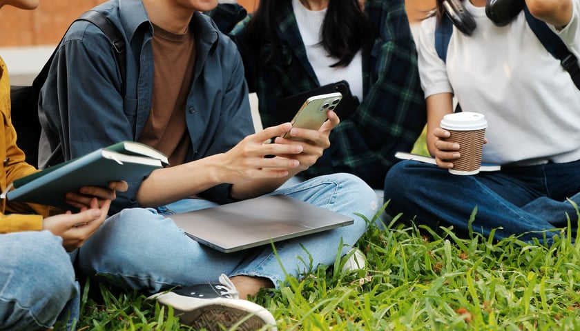 Grupa studentów siedząca na trawie. Jedna osoba trzyma smartfon i ma laptop na kolanach, inna trzyma książki, kolejna kawę w papierowym kubku. Wszyscy mają plecaki i słuchawki.