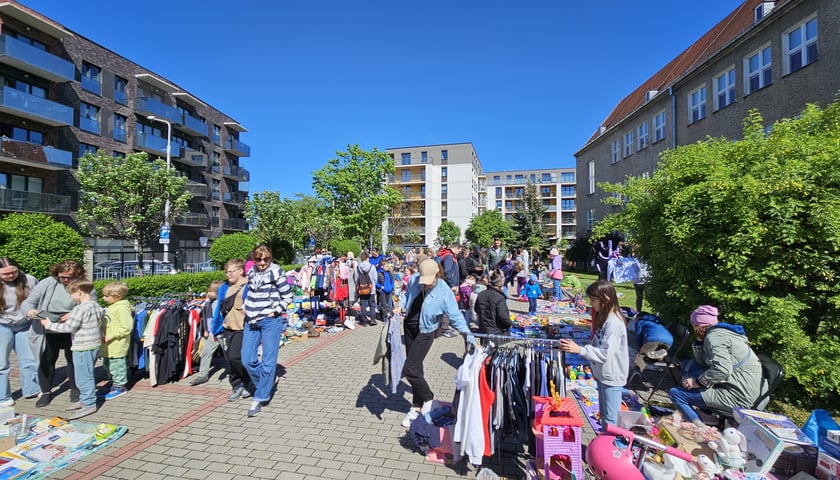 Widoczna uliczka osiedlowa na Tarnogaju. Po obu stronach widoczne punkty z różnymi rzeczami na wymianę, wśród których spacerują mieszkańcy osiedla.