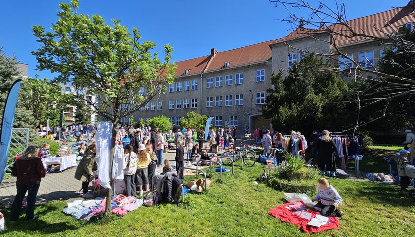 Po lewej stronie widać wieszaki z kolorowymi ubraniami, a po lewej stronie, na zielonym trawniku siedzi młoda dziewczyna na czerwonym kocu i rozkłada materiały do rękodzieła.
