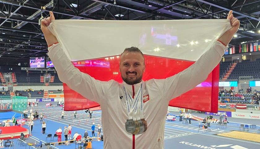 Wrocławianin Andrzej Kominek stoi z polską flagą na podium z czterema medalami na szyi: brązowym w rzucie młotem, srebrnym w pchnięciu kulą i rzucie ciężarkiem i złotym w rzucie dyskiem. 