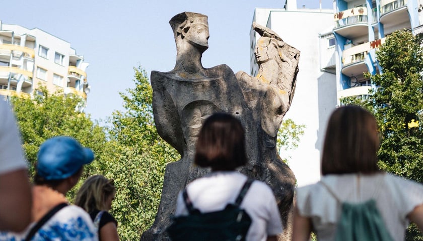 Sąsiedzki spacer po osiedlu z Mikrograntami. Zdjęcie archiwalne. Na zdjęciu grupka osób stojących przy pomniku., W tle bloki i drzewa.