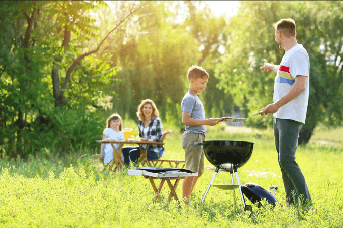 Tata i syn stoją nad otwartym okrągłym grillem ogrodowym na trawniku, w tle mama z córką siedzą przy stoliku