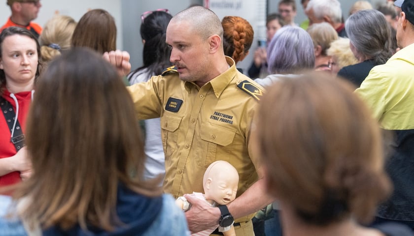 Mężczyzna w mundurze Państwowej Straży Pożarnej rozmawia z osobami otaczającymi go kołem, w lewej ręce podtrzymując fantom niemowlęcia