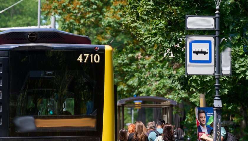 Zdjęcie ilustracyjne - tablica przystanku autobusowego i fragment autobusu na tle drzew.