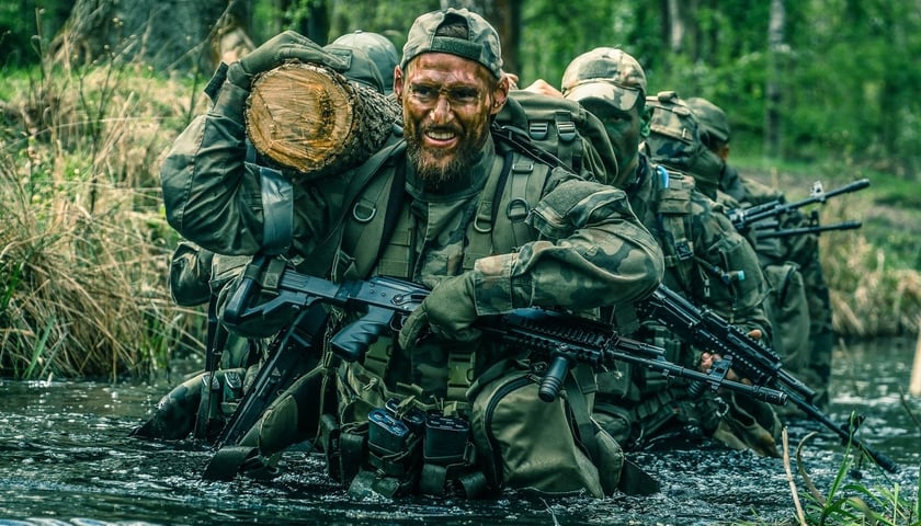 Mężczyzna  w  wojskowym mundurze polowym w rzece, niesie pień drzewa, ma przewieszony karabin, w tle inni żołnierze. Uczestnicy kursu Piorun w 2026 r. 