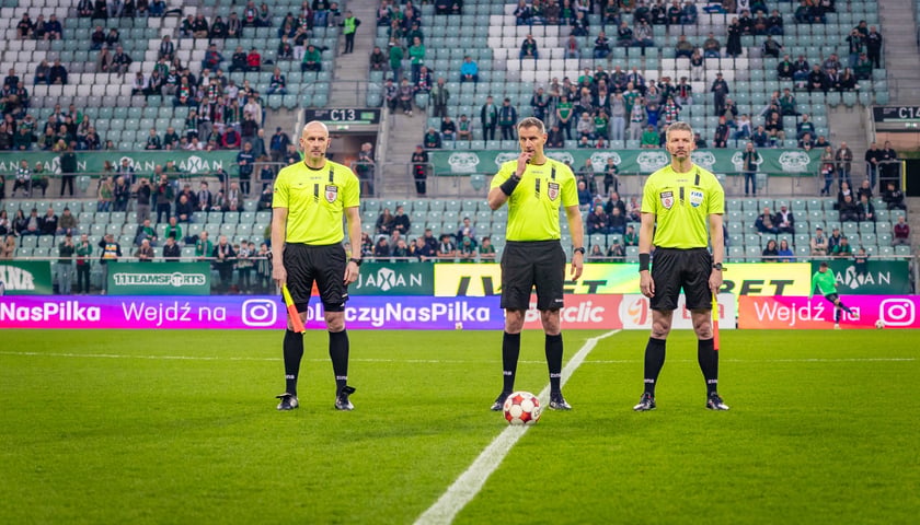 Trzech sędziów stojących na środku boiska z piłką, w tle trybuny stadionu częściowo puste. Mecz Śląsk Wrocław - Wisła Kraków 7 marca 2026 r. na Tarczyński Arena zakończony walkowerem z powodu nieobecności Wisły Kraków.