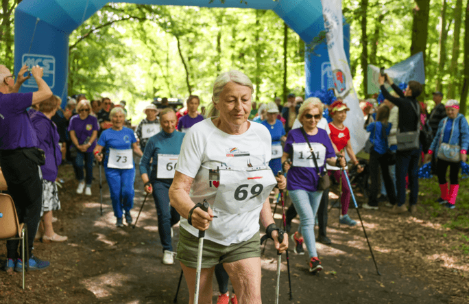 Siwowłosa kobieta w jasnej koszulce z numerem startowym 69 i krótkich spodenkach. Maszeruje, trzymając kijki do nordic walking