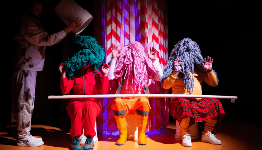 Zdjęcie z teatru. Obok siebie siedzą trzy postacie w wielkich czuprynach z frędzli (zielona, różowa, szara) przypominających mopy. Po lewej stoi mężczyzna w jasnym stroju i trzyma białe wiadro nad zielonowłosą postacią. Scena jest ciemna i kolorowa