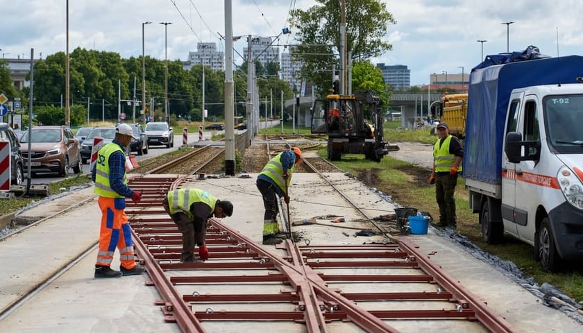 Robotnicy w kamizelkach odblaskowych montujący nowe torowisko tramwajowe na betonowym podłożu w sąsiedztwie ruchu ulicznego i pojazdów technicznych