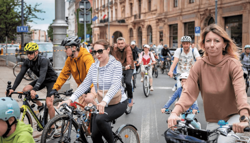 Duża grupa rowerzystów w różnym wieku, jadących kolumną przez miejską ulicę wzdłuż zabytkowych kamienic