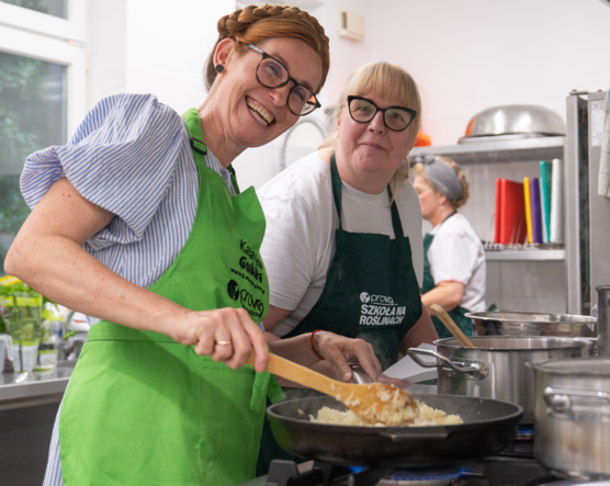 Dwie uśmiechnięte kobiety w okularach i fartuchach kuchennych. Ta z lewej to Katarzyna Gubała - miesza potrawę na patelni drewnianą łyżką podczas warsztatów kulinarnych w profesjonalnej kuchni