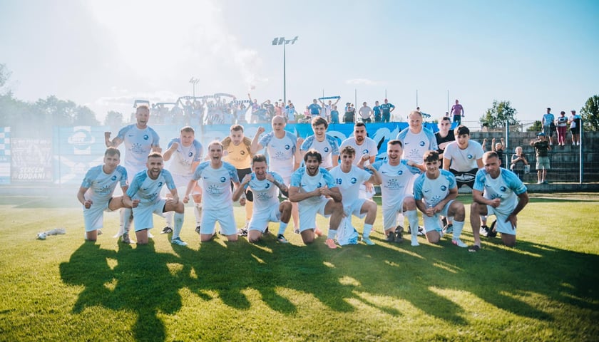 Piłkarze Polaru Wrocław po meczu z Pogonią Oleśnica na stadionie na wrocławskim Zakrzowie. Za nimi na trybunach wierni kibice z biało-niebieskimi szalikami.