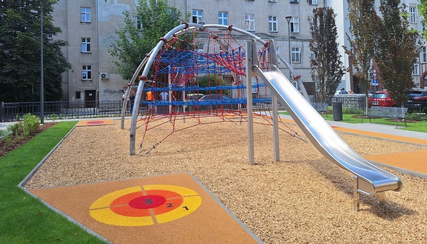 Plac zabaw na którym widać urządzenie wielofunkcyjne do wspinaczki, zjeżdżalnie i stałą grę podwórkową - rzut do celu.