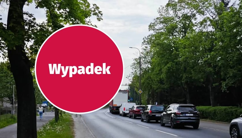 W tle - droga i stojące na jednym pasie samochody. Zdjęcie ilustracyjne. W kółku - czerwona plansza z napisem "wypadek".