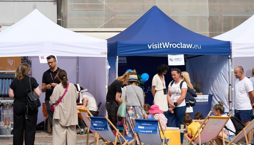 Dzieci trzymające niebieskie baloniki smartcity Wrocław przy granatowym namiocie z napisem "visitWroclaw.eu", w tle stoisko z rollupem miasta i uczestnicy wydarzenia.