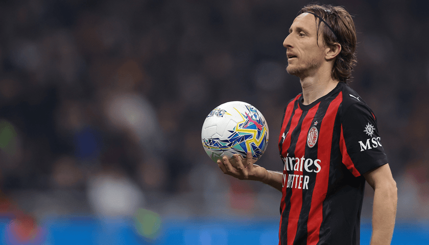 Luka Modrić w czarno-czerwonym stroju Milanu, trzymający piłkę na dłoni