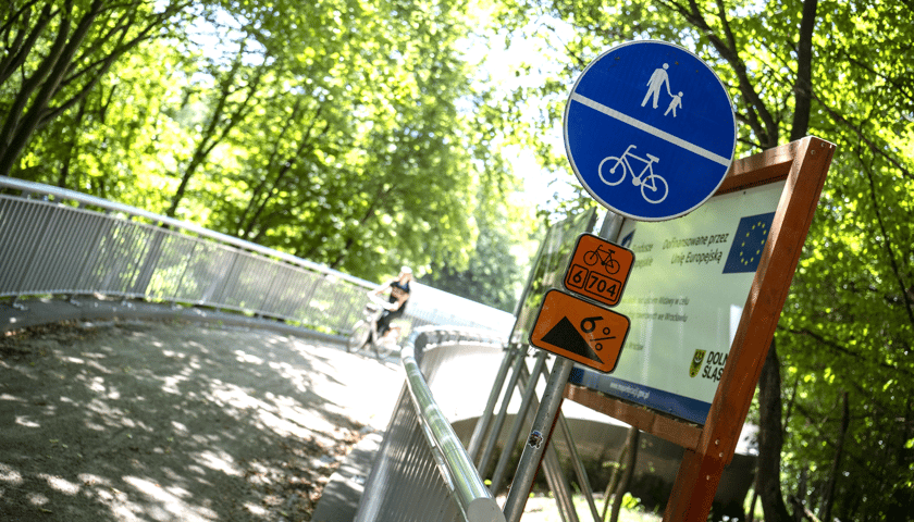Wjazd na kładkę nad Widawą - przy barierach znaki z numerem trasy rowerowej, informacja o nachyleniu i znak o drodze pieszo-rowerowej, w tle widoczny rowerzysta i drzewa.
