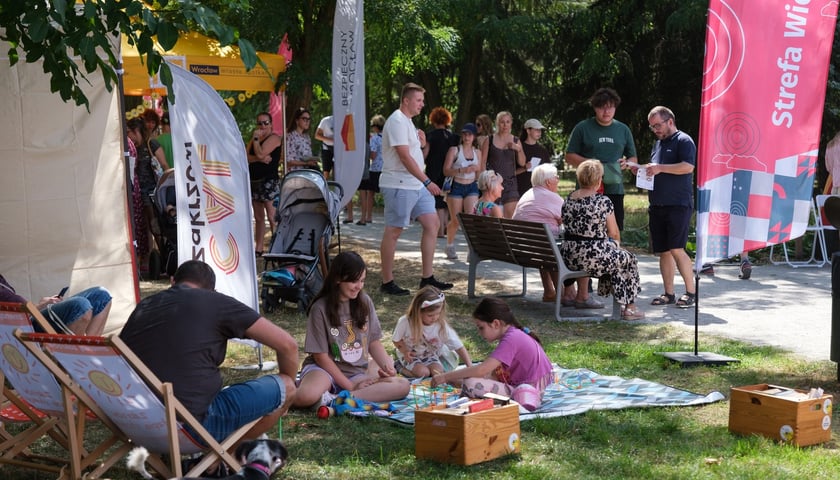 Zdjęcie przedstawia uczestników plenerowego pikniku portalu wroclaw.pl w parku, gdzie na kocu dzieci układają klocki, a w tle dorośli rozmawiają siedząc i stojąc