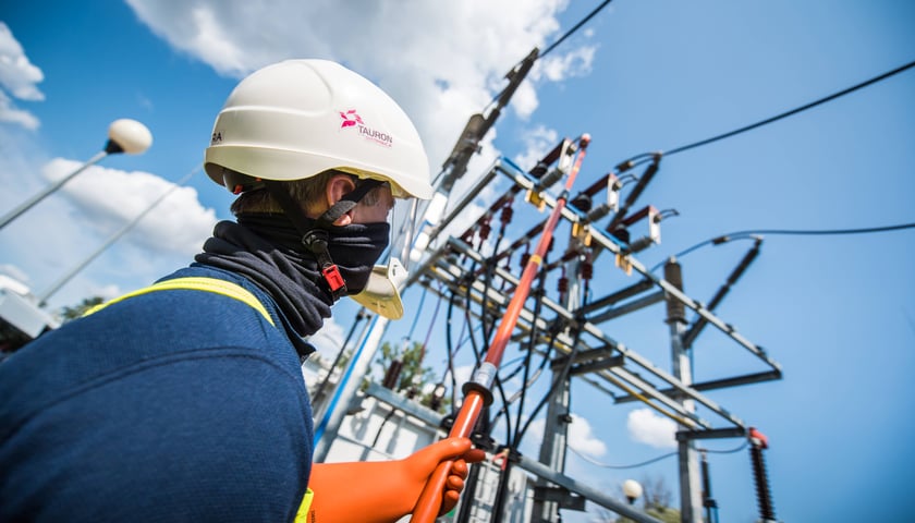 Elektryk w białym kasku i osłonie twarzy, z logo Tauron, obsługuje pomarańczowym drążkiem izolacyjnym aparaturę na stacji elektroenergetycznej. 18 sierpnia 2026 r. doszło do awarii prądu we Wrocławiu