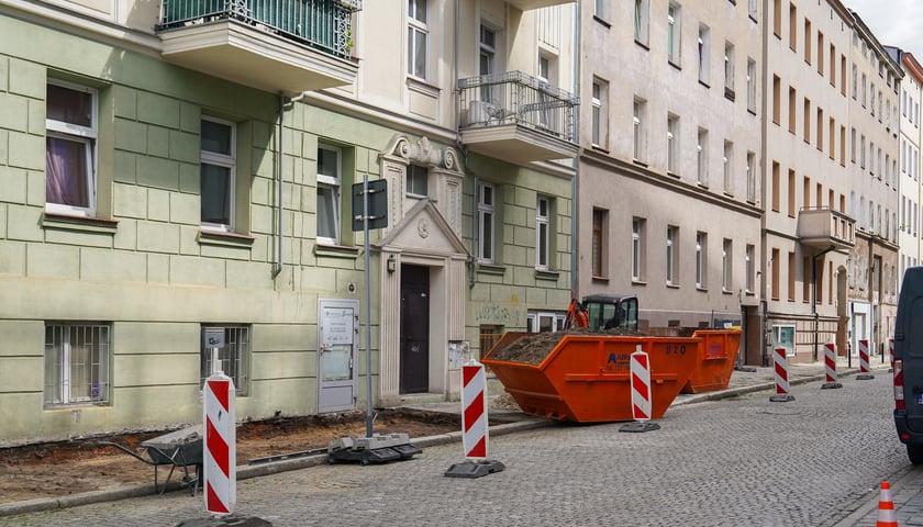 Słupki drogowe i kontenery na gruz stojące na brukowanej jezdni Pl. Konstytucji 3 Maja przy krawędzi chodnika, który jest remontowany.