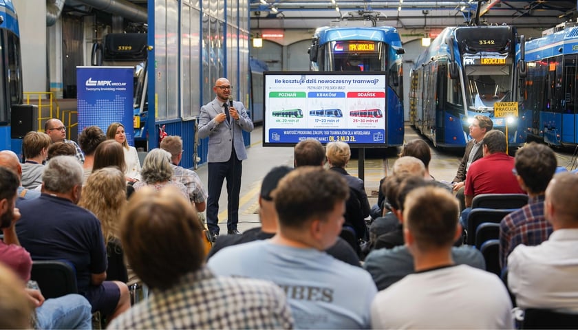 Prezes MPK Wrocław Witold Woźny na spotkaniu z mieszkańcami. W tle widoczne są różne modele tramwajów, koło prezesa stoi tablica z tytułem "Ile kosztuje dziś nowoczesny tramwaj?"