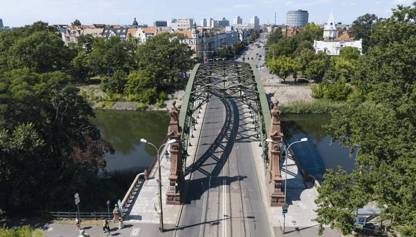 Zdjęcie z lotu ptaka pokazuje zielony, stalowy most z torami tramwajowymi nad rzeką. Przy wjazdach stoją wieżyczki, wokół rośnie dużo zieleni, a w tle widać Wrocław. Most Zwierzyniecki znalazł się na liście, ogłoszonej latem 2026 przez Zarząd Dróg i Utrzymania Miasta, programu remontów mostów. 