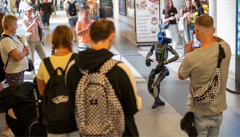 Humanoidalny robot "Adam Kowalski" tańczy przy wejściu na perony dworca Wrocław Główny. Występowi przygląda się tłum ludzi, kilkoro z nich robi zdjęcia telefonami komórkowymi.