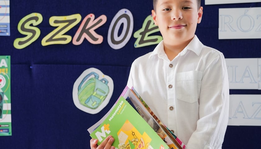 Franek, uczeń wrocławskiej podstawówki stoi w białej koszuli i trzyma książki przed napisem "Witaj szkoło"