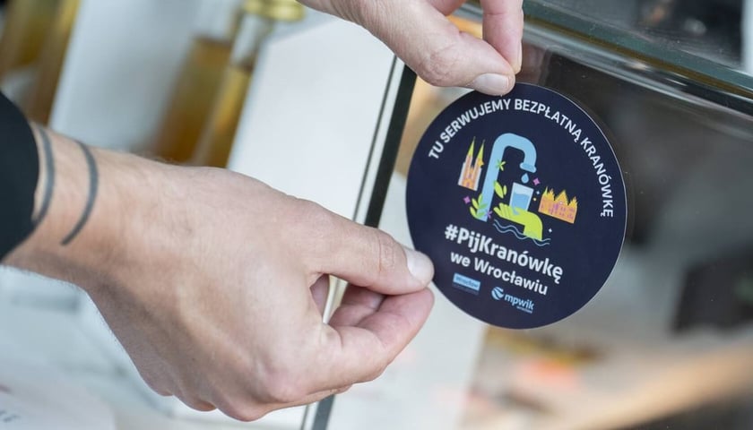Руки наклеюють наліпку, яка повідомляє, що в цьому закладі подають воду з-під крана