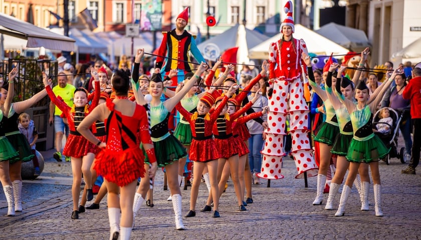 Grupa młodych tancerek w strojach estradowych maszeruje w radosnej paradzie na wrocławskim Rynku w towarzystwie szczudlarzy przebranych za krasnale